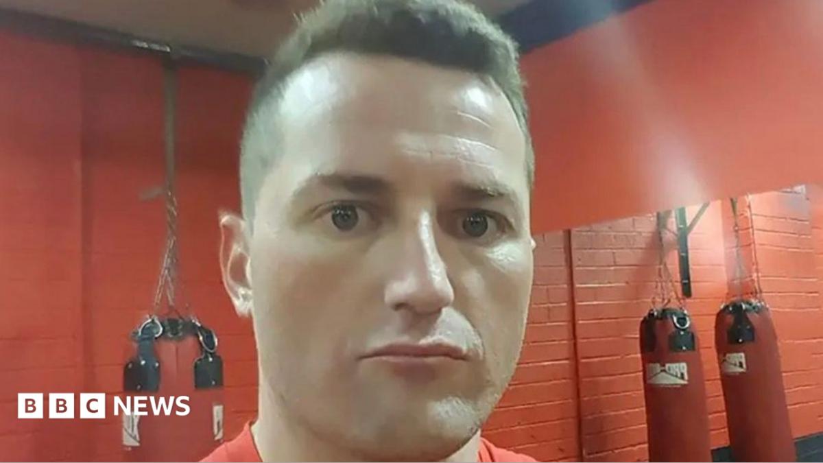 Robbie Lawlor. He has short brown hair and brown eyes, wearing a red t-shirt. There is red brick walls, a mirror and boxing bags behind him.