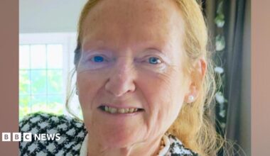 A woman in her late 60s, with blue eyes and light ginger hair tied back, smiles at the camera. She is inside a house with a green curtain and window behind her, and is wearing a black and white chequered jacket.