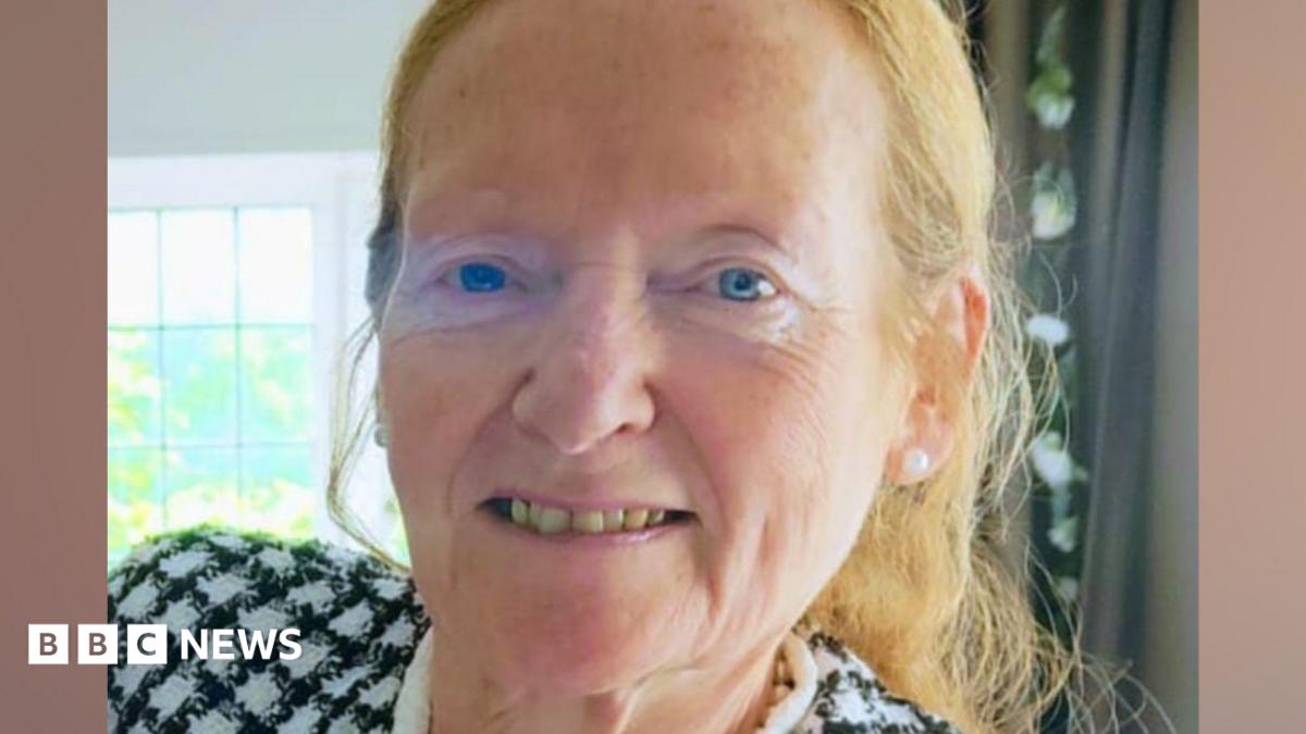 A woman in her late 60s, with blue eyes and light ginger hair tied back, smiles at the camera. She is inside a house with a green curtain and window behind her, and is wearing a black and white chequered jacket.