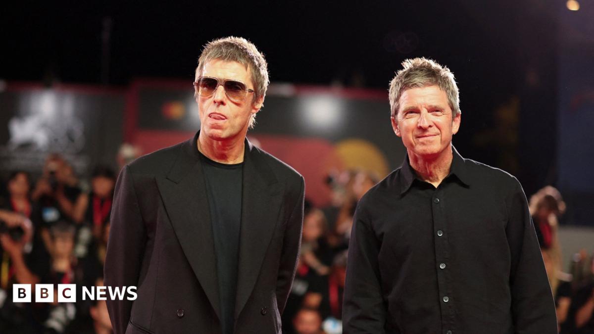 Liam and Noel Gallagher wearing black clothing stand on a red carpet with photographers stood behind them.