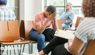 A stock image of people sat in a hospital waiting room. One woman with short dark hair, wearing a pink t shirt, has her head in her hands.