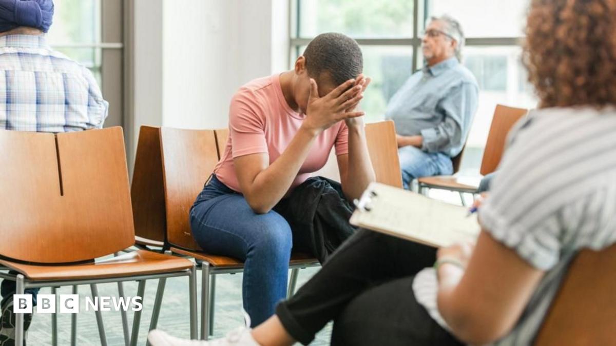 A stock image of people sat in a hospital waiting room. One woman with short dark hair, wearing a pink t shirt, has her head in her hands.