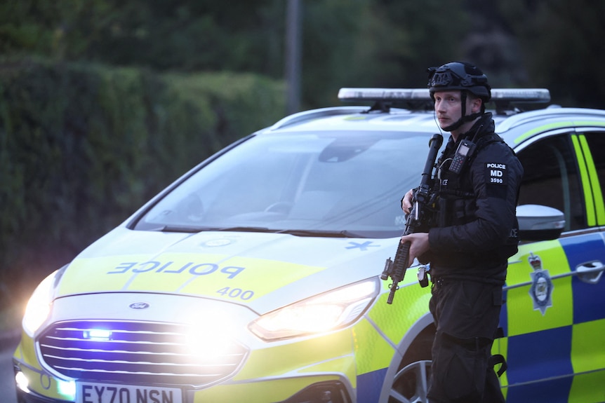 A British counterterror policeman wearing black clothes and helmet while holding a rifle, in front of a blue and yellow car.