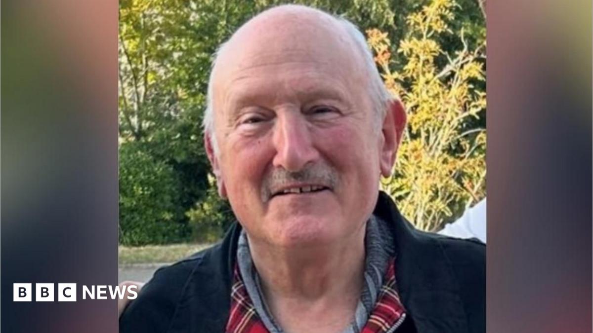 A man with greying short hair and a short moustache. He is wearing a black jacket with red tartan lining and a shirt underneath. He is smiling. Trees are behind him.