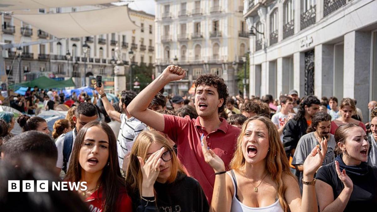 Four women and a man shout and raise their arms while protesting at Puerta del Sol in Madrid. Crowds of other people stand around them.