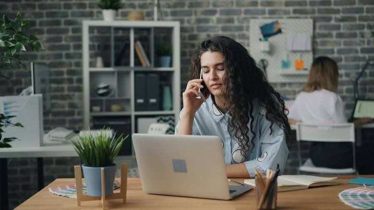 A woman is on the phone while she sits at her desk in an office; another person in the background is sitting at another desk with their back to her.