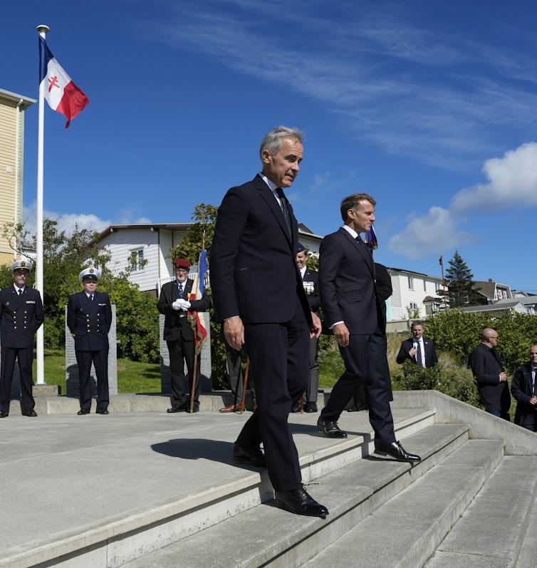 Two men in suits walk down a short flight of stairs as a French flag flies behind them on a sunny day.