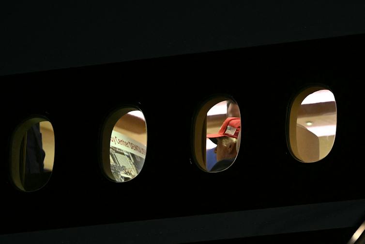 A man in a baseball cap is seen aboard an airplane, apparently reading the words 'Kennedy Center demolished.'