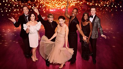 Strictly judges Craig, Shirley, Motsi and Anton strike a celebratory pose alongside new hosts Josh, Emma and Johannes in a ballroom lit by glowing golden lights. The women wear dresses and the men wear dark outfits.