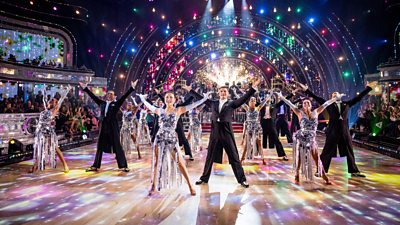 The Strictly pro dancers stand on the ballroom floor, arms raised in black suits and silver dresses