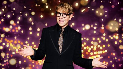 Josh Widdicombe stands with his hands out smiling to camera in a dark suit and patterned tie, set against a dark background dotted with warm gold lights