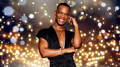 A man in a black outfit, cut with a deep v at the chest, smiles to camera