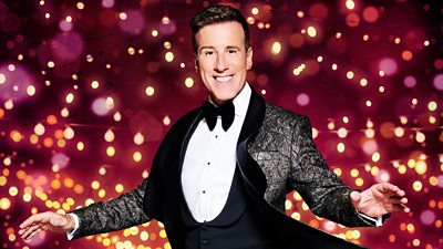 A man in a patterned suit jacket, white shirt and bow tie smiles to camera as he poses with his arms out, ready to dance