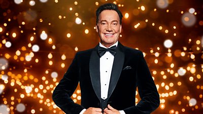 A man in a black suit, bow tie and white shirt smiles to camera as he closes his jacket