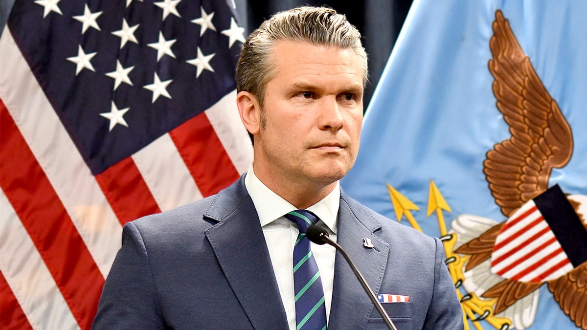 War Secretary Pete Hegseth looks on with flag behind him.
