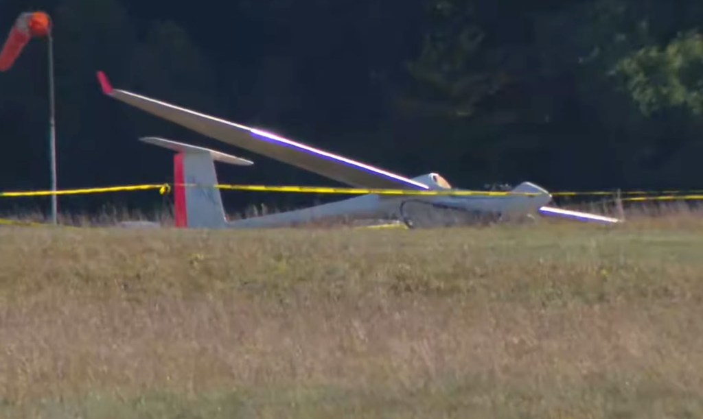 A crashed glider plane behind yellow caution tape on a grassy field.
