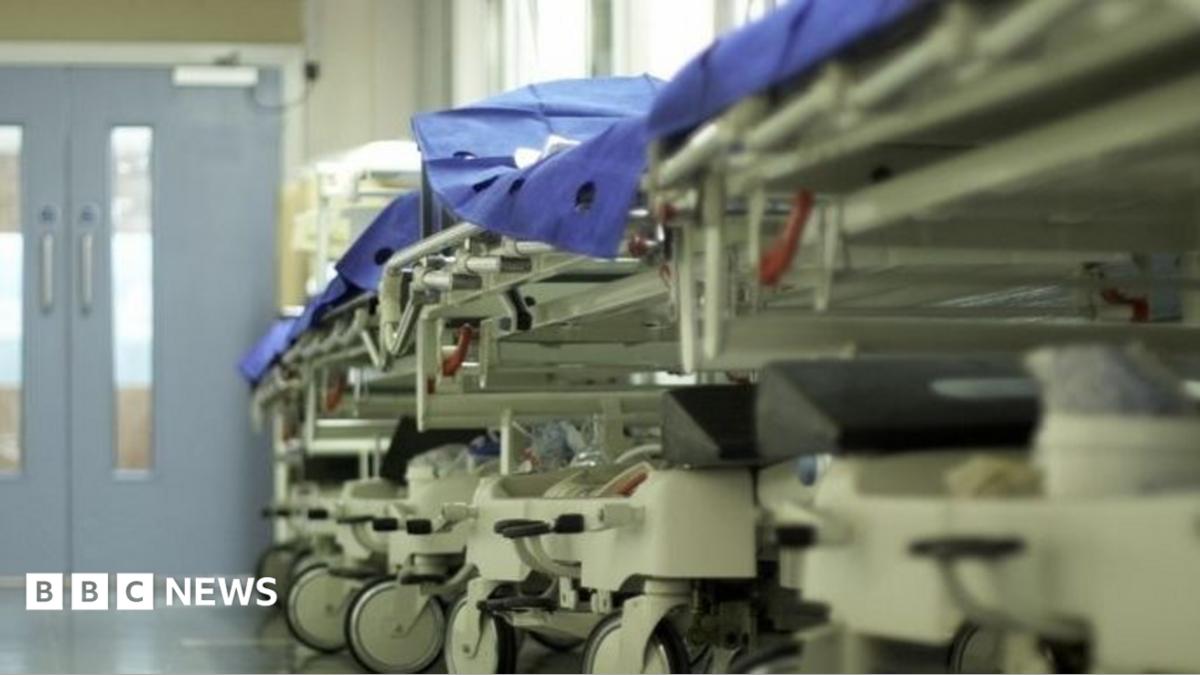 A row of empty hospital trolleys line a brightly lit corridor, their wheels reflecting on the polished floor. Blue protective sheets cover the beds, while there are closed double doors at the far end