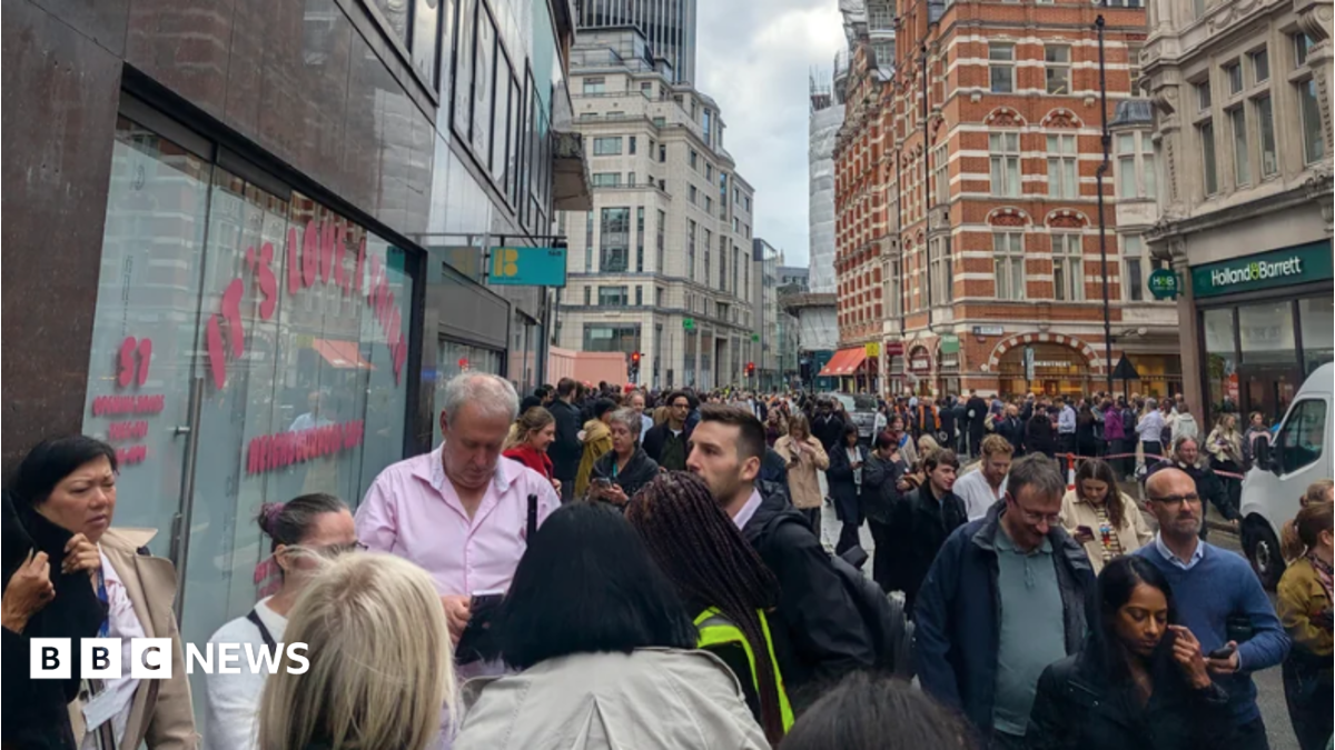 Crowds of people stand on a pavement and spill into the road on a street in the City of London, surrounded by office buildings and shops.