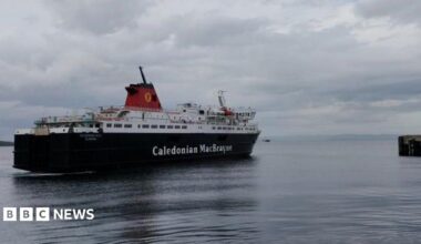 MV Caledonian Isles sailing across a calm harbour toward a pier on the right, beneath a cloudy grey sky. The black-and-white vessel is reflected in the still water, with distant coastline visible on the horizon.