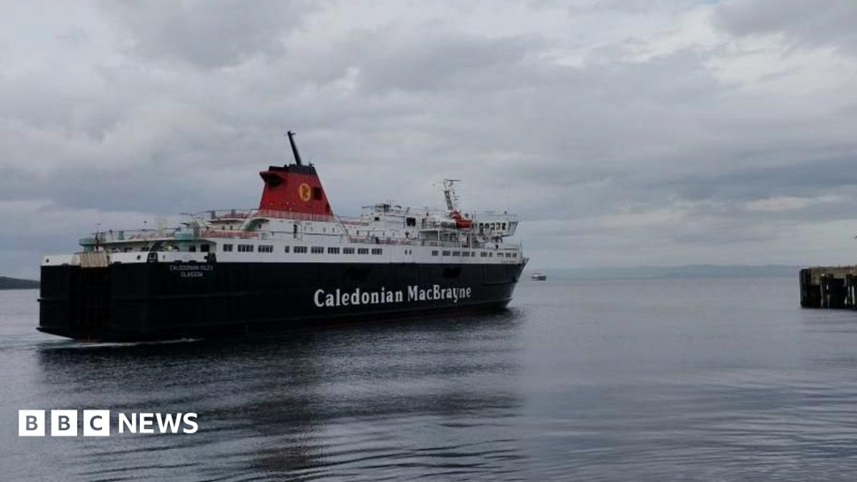 MV Caledonian Isles sailing across a calm harbour toward a pier on the right, beneath a cloudy grey sky. The black-and-white vessel is reflected in the still water, with distant coastline visible on the horizon.