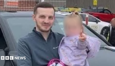 Cody Lowndes standing beside a dark SUV in a parking area, holding a small child whose face is blurred. The child is wearing a light purple top. A metal fence, several parked vehicles and an industrial-style building are visible in the background. The image appears to have been taken outdoors during daylight.