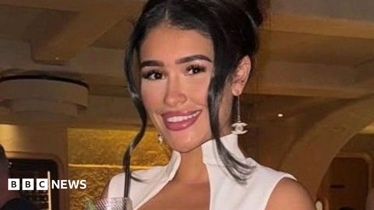 Alisha Samuels has long brown hair and is wearing ear rings and a white dress.