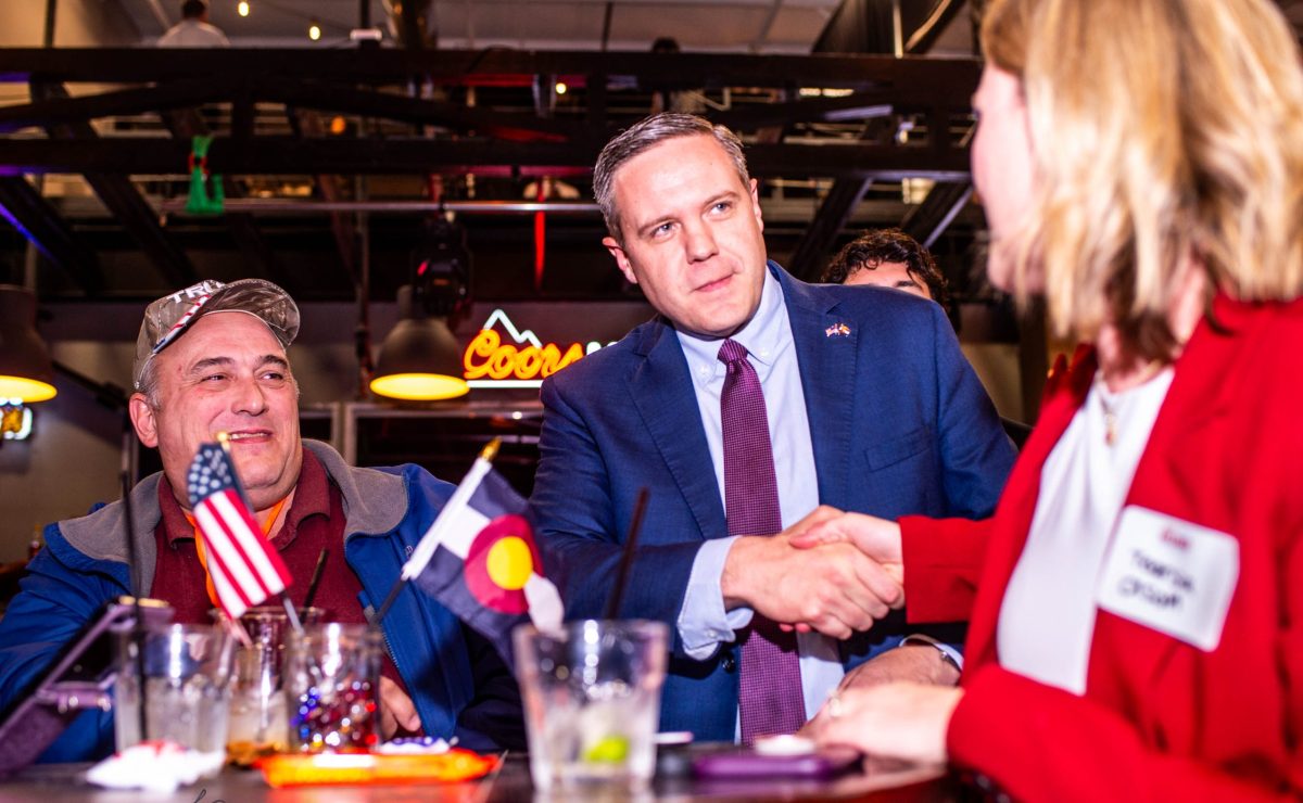 A man in a blue suit and purple tie, with a flag pin on his lapel speaks with enthusiastic supporters at his watch party