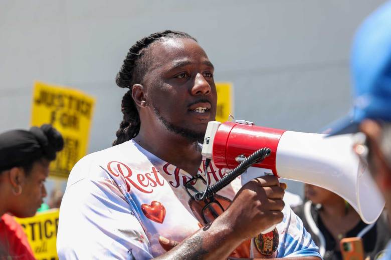 A man wearing a "Rest in Heaven" tshirt holds a white megaphone at a gathering