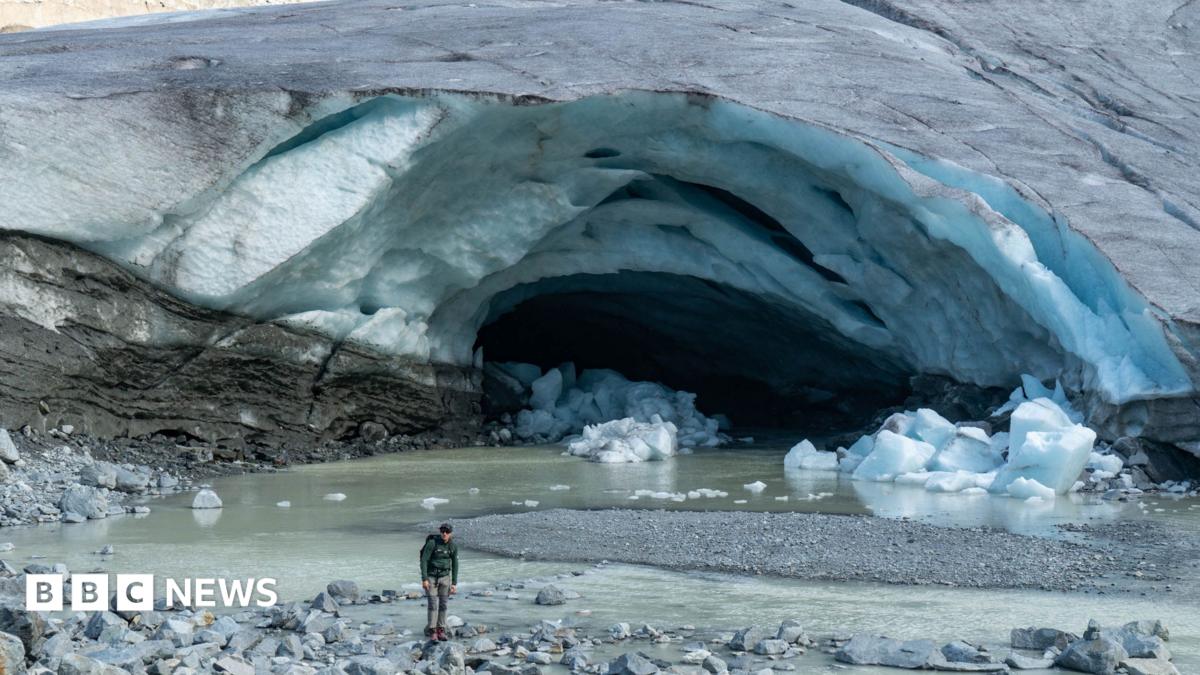 The front of a glacier, where a large channel - which looks a bit like a cave or an arch - has eaten away at the ice. A river of meltwater now meanders out of the front of the glacier. There are also chunks of ice on the ground having broken off the glacier. A man, dressed in mountaineering clothing, stands in front of the glacier for scale, which shows how big the channel or cave is.