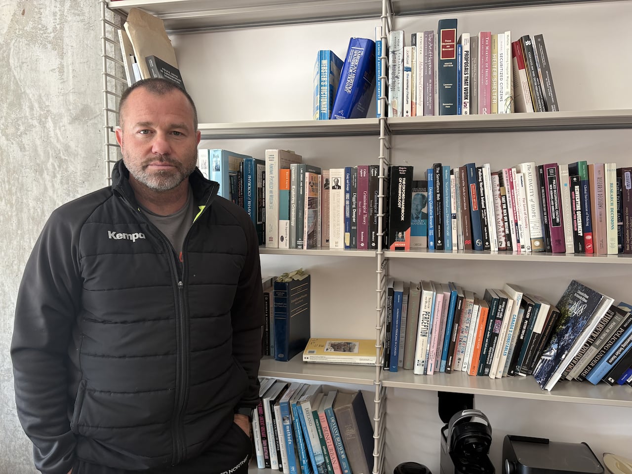 A white man with a buzz cut stands next to a book shelf and looks at the camera.