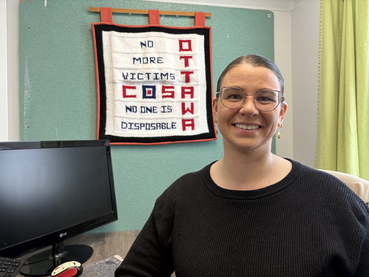 A woman in a black sweater smiles at the camera. She is sitting at a desk and behind her there is a tapestry that reads "no more victims, no one is disposable, CoSA Ottawa."