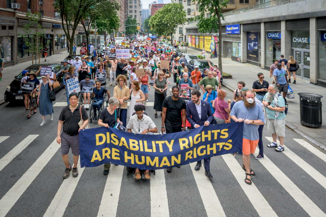 A big crowd of people is marching down a street in New York holding up a banner that says disability rights are human rights in big yellow letters on a blue background.