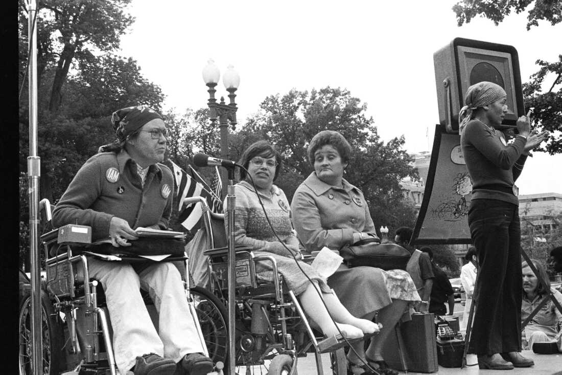 A black and white image of activist Kitty Cone speaking into a microphone with Judy Heumann and Eunice Fiorito sitting by her side on a stage, speaking to a crowd. Sign language interpreter Jadin Murello is standing at the end of the row, signing to the audience.