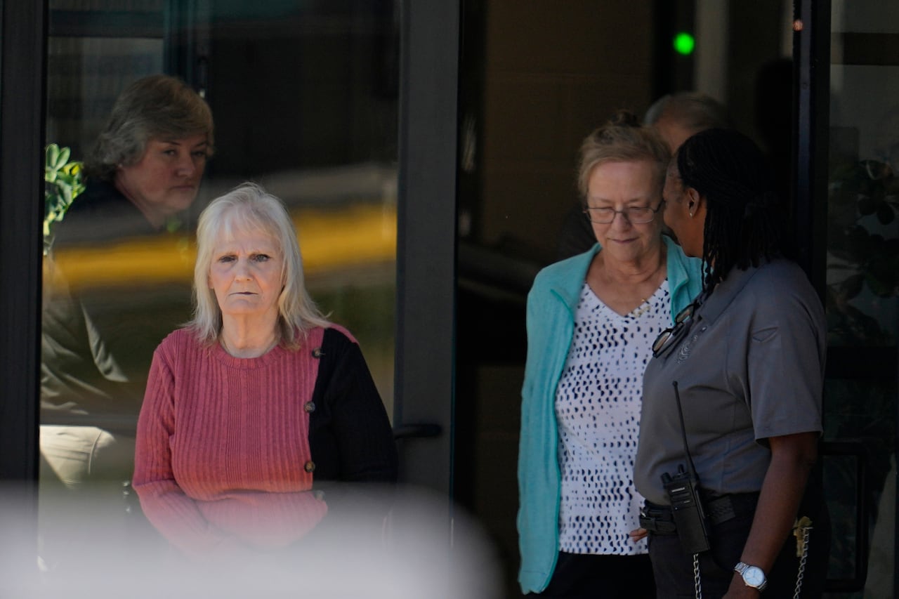 Two older women walk outside a building. The woman on the left wears a rose-coloured sweater. The woman on the right wears a light blue cardigan over a blouse.