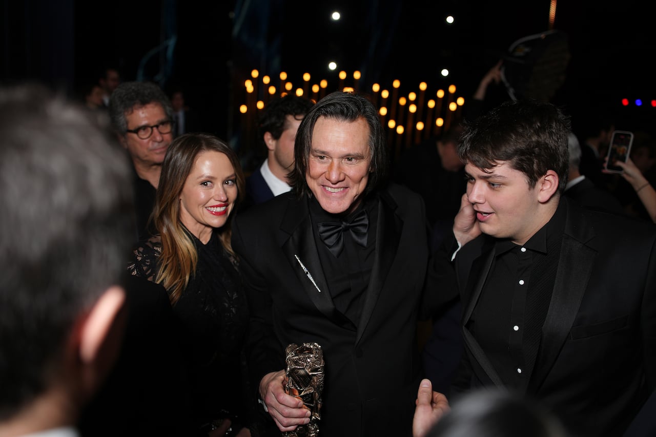 A man holding a gold award poses for photos alongside a woman and young man dressed in black. Lights shine behind them.