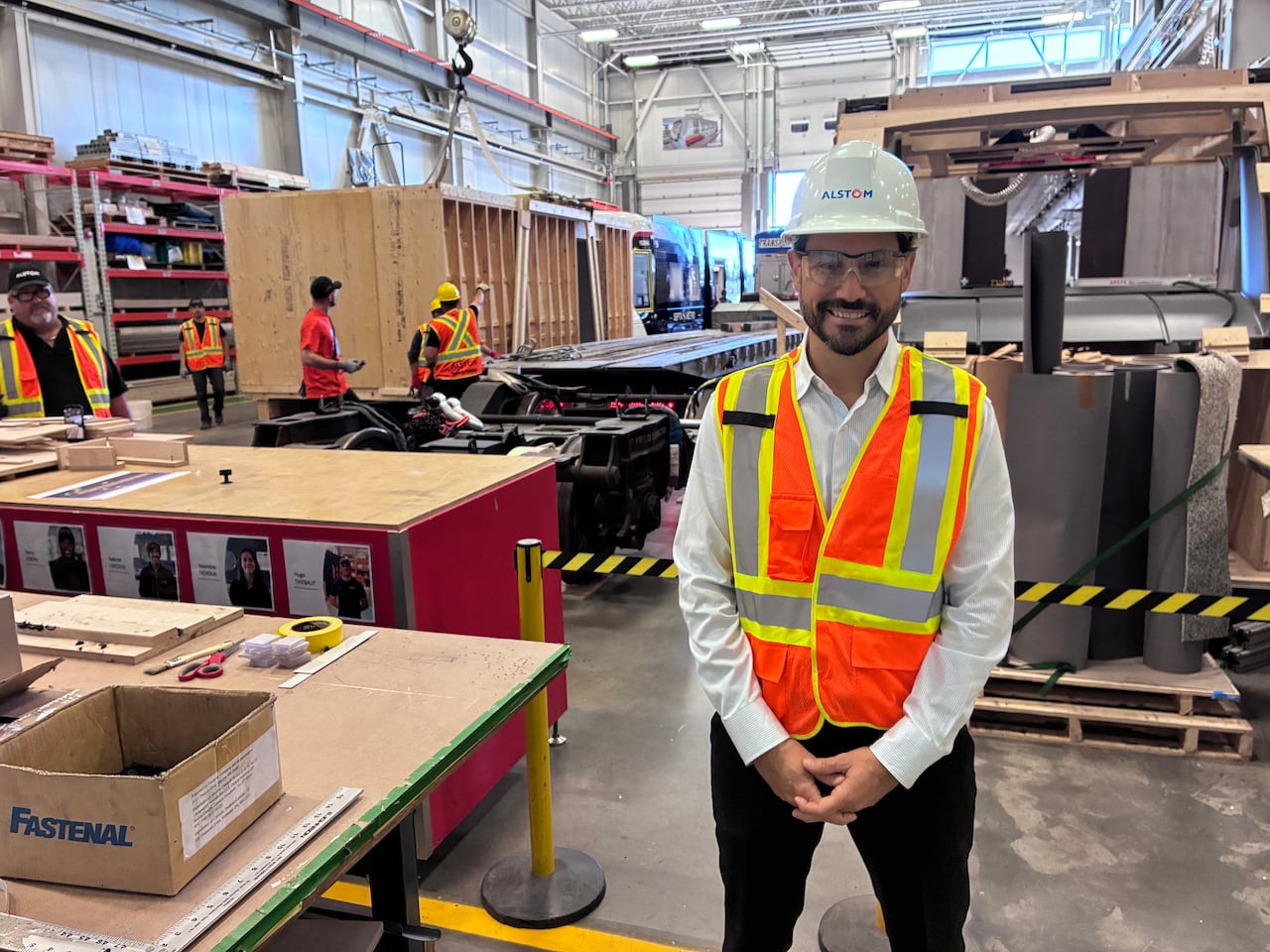 A man in an orange safety vest, goggles and a hard hat stands on a factory floor.