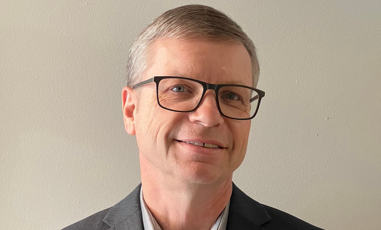 Man with grey hair wearing black square glasses and a grey suit smiles at the camera.