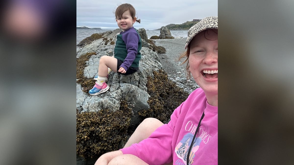 A toddler sitting on a rock on a beach with her mom partially in the photo as well.