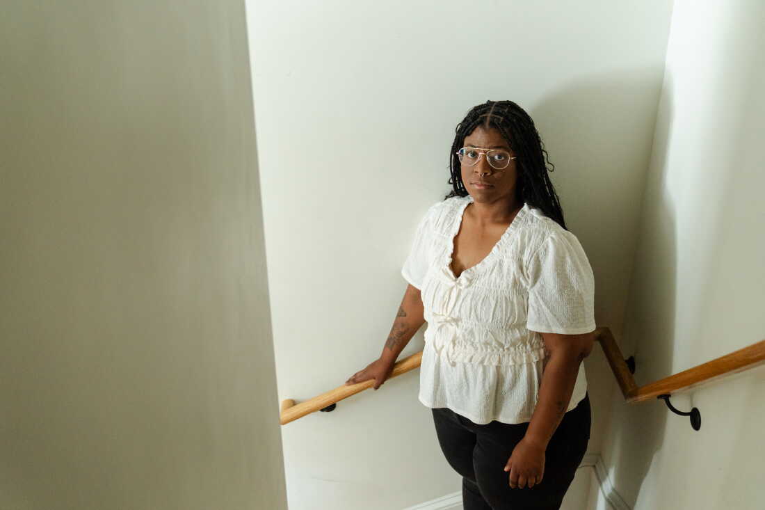 Marissa Pike has long, dark braids, has glasses and is wearing a white top. She is standing on a landing in her home.