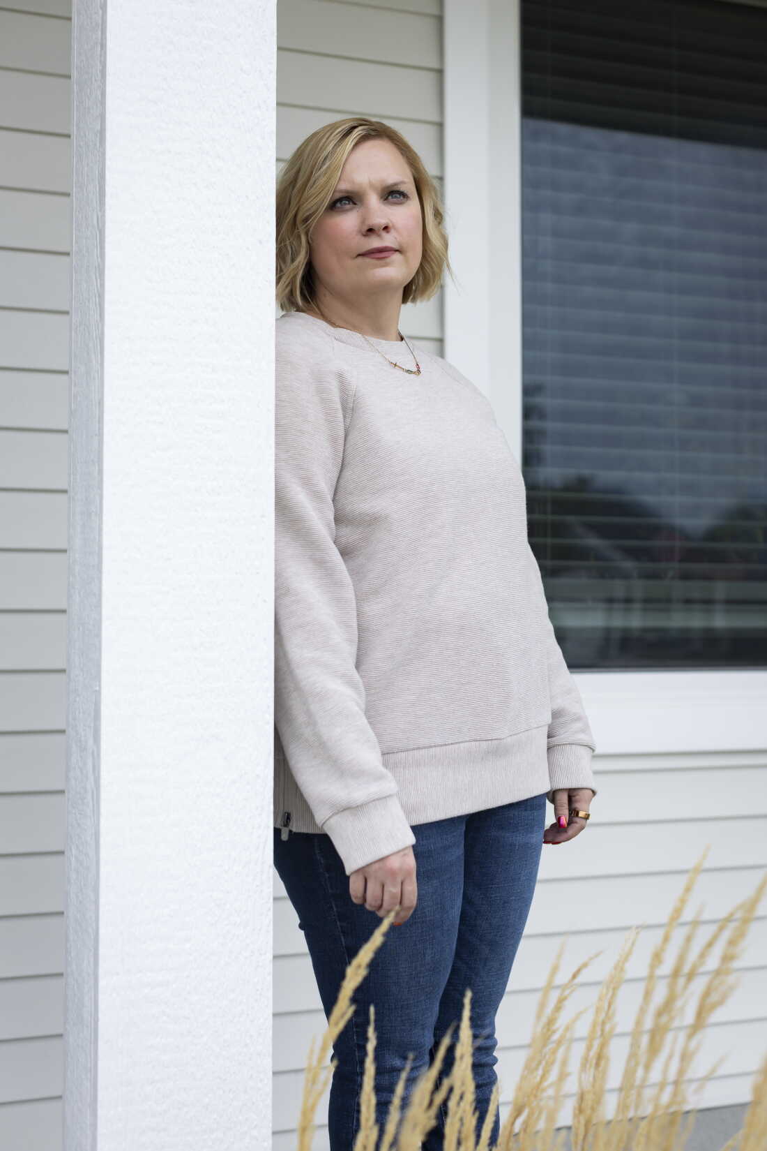 Samanthya Marlatt, who has cerebral palsy and spent time in foster care, lives in Iowa. She has chin-length blonde wavy hair and is wearing a cream colored sweater while standing outside her home.