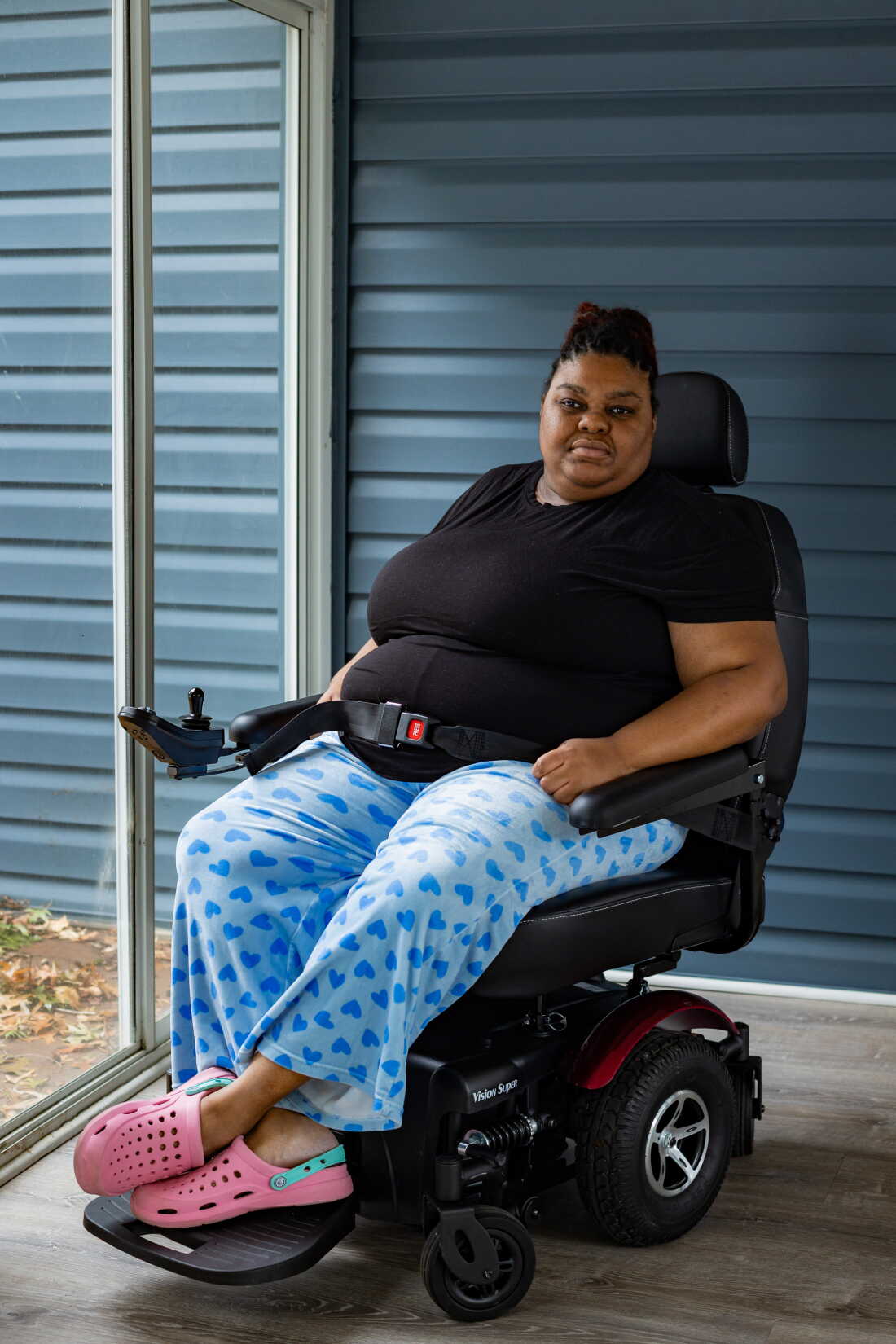 Rasheeda McCrae, 35, who is legally blind, uses an electric wheelchair and is sitting on the back porch at her home. She has dark braids and is wearing a black t-shirt with blue patterned pants and pink rubber shoes.