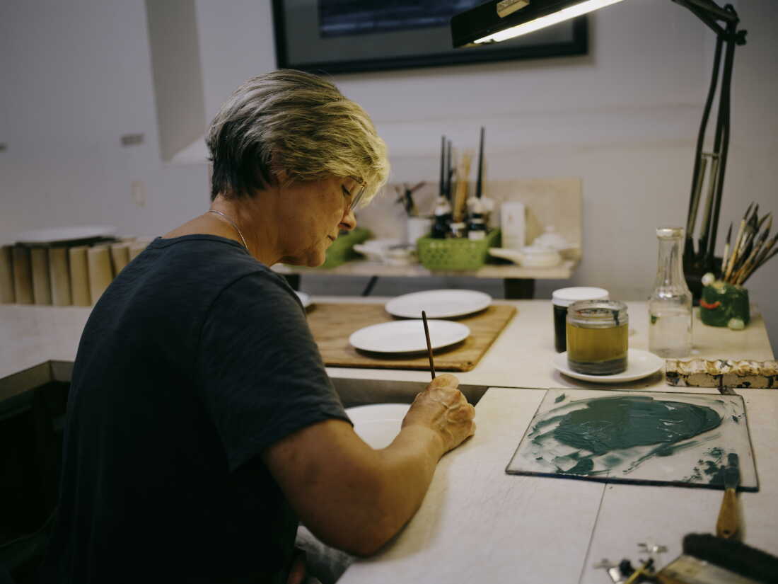 A blonde woman dips a paintbrush into green paint and paints a small white china plate.
