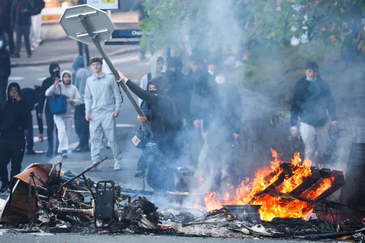 Several people, some wearing masks, are shown in an apparent outdoor demonstration. Some items are on the ground burning, while one person holds up a street sign.