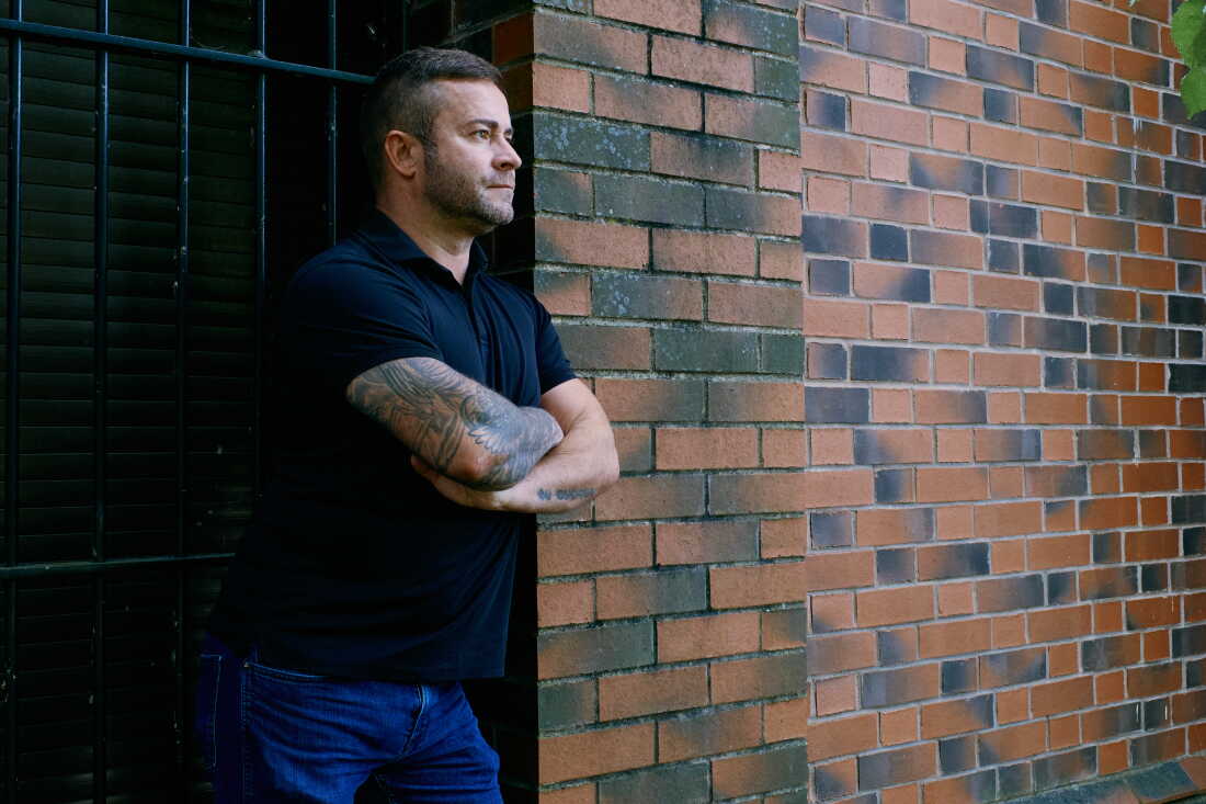 Shane Thomson stands next to a brick wall. He's wearing jeans and a navy blue short-sleeved shirt.