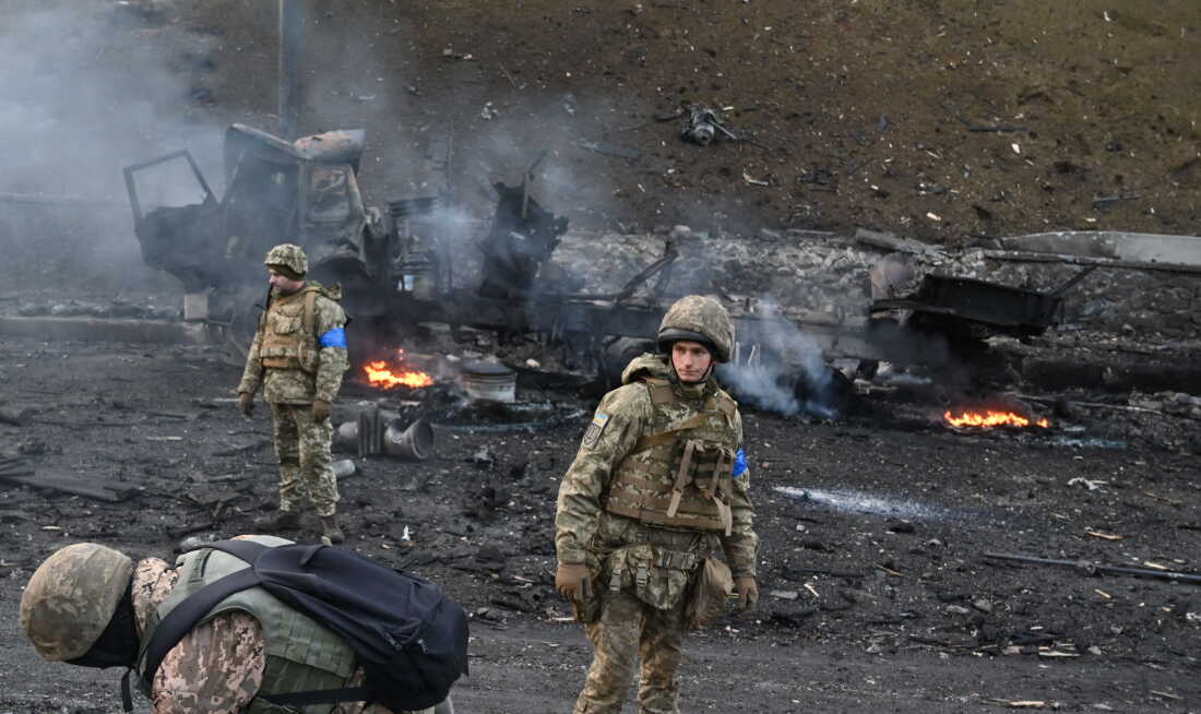 Three Ukrainian service members in camouflage uniforms look for and collect unexploded shells in Kyiv on February 26, 2022. Small fires are behind them among the rubble.