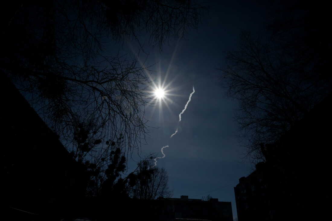 A white rocket trail appears against a dark blue sky over Irpin on March 12, 2022. Tree branches are silhouetted against the dark sky.