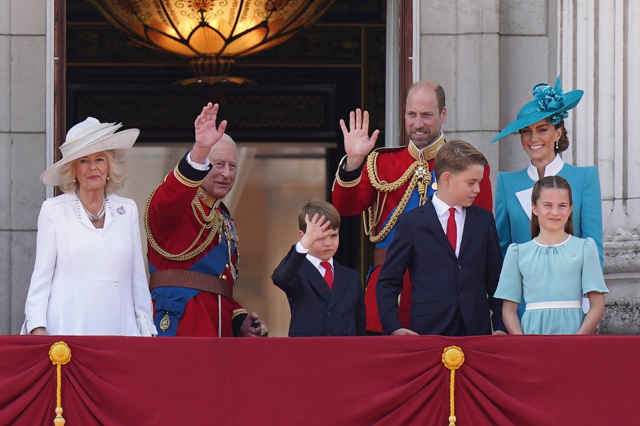 Four adults and three children wave as they stand on the balcony of a large stone building.