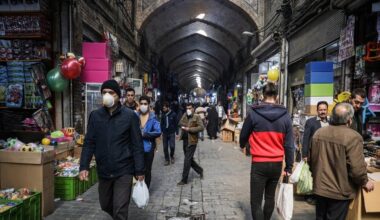 A quiet current of daily life moves through the vaulted corridors of an old covered bazaar, where shoppers weave past toy stalls, hanging balloons, and stacked crates under rows of hanging lamps.
