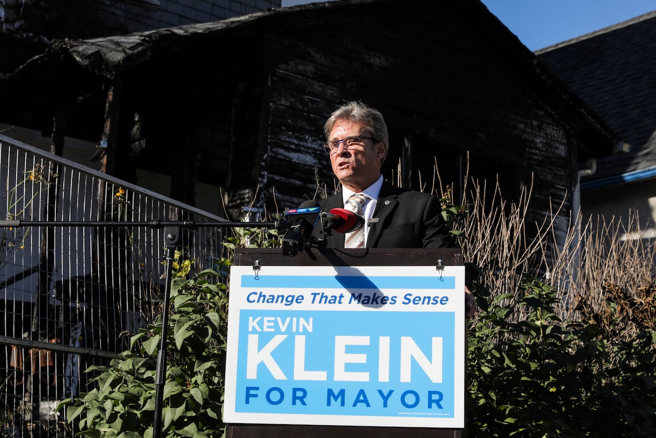 A man stands at a podium in front of a burned-out house.