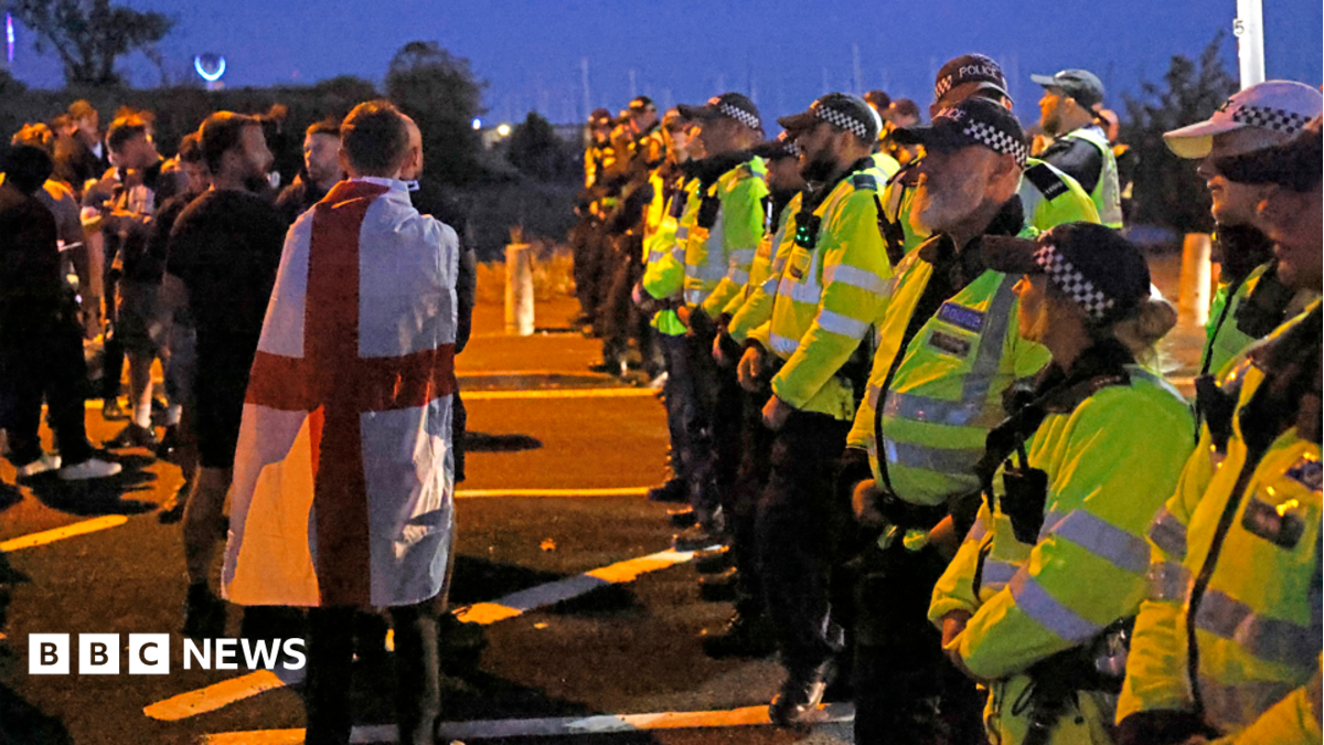 There is a line of police to the right of the image, facing towards the left. They're wearing yellow hi-vis jackets and black and white hats. On the left of the image are a handful of men, one wrapped in a red and white flag